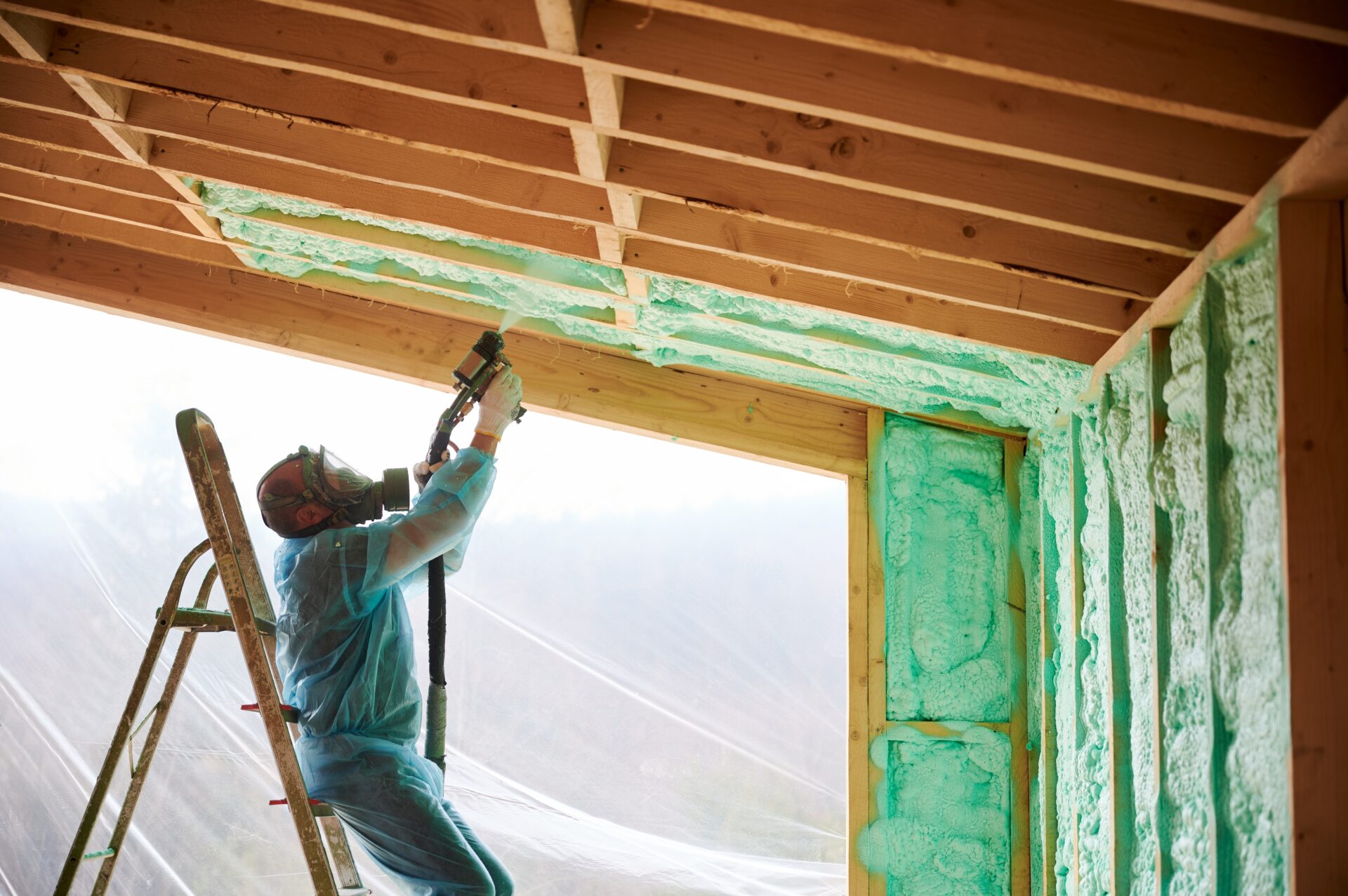 A person wearing blue coveralls and a respirator mask installs spray foam insulation into a ceiling panel