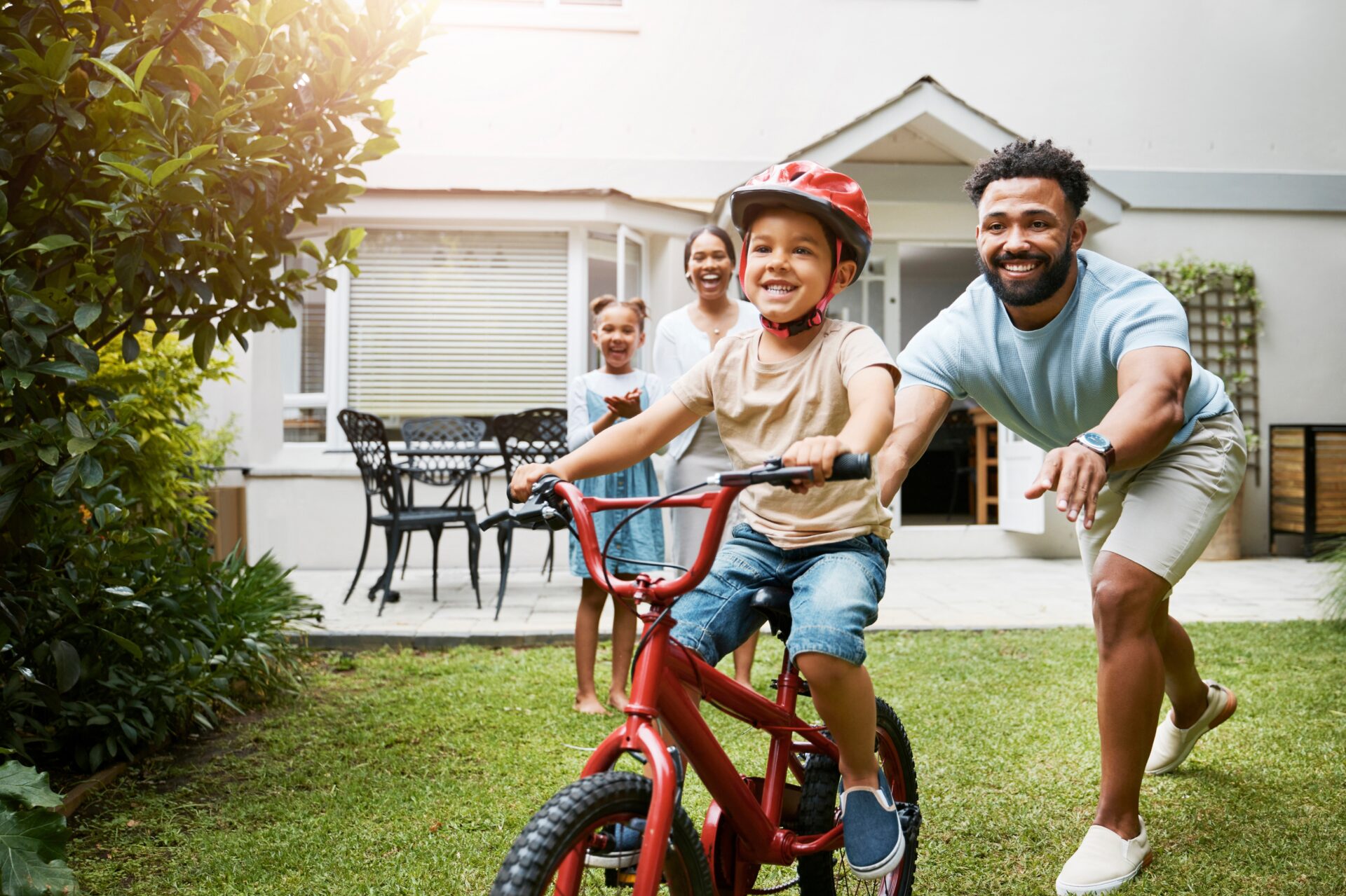 A man in shorts teaches his young son to ride a red bicycle in a home’s backyard while his wife and daughter watch and clap 