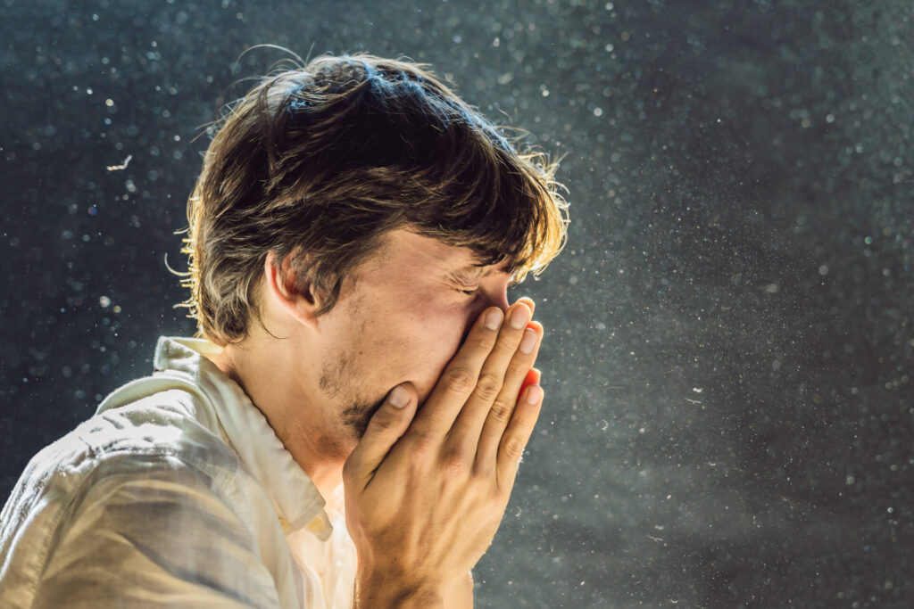 A man sneezes into his hands with visible particulate matter in the air