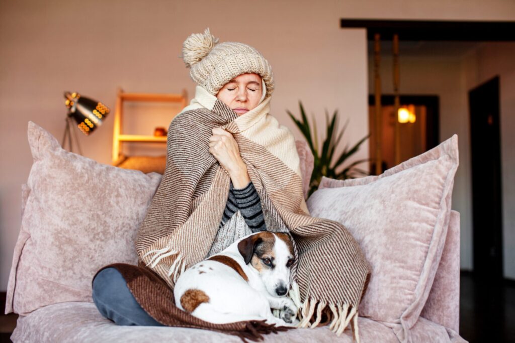 A woman sits on a sofa wearing a knitted hat and wrapped in a blanket with a dog in her lap
